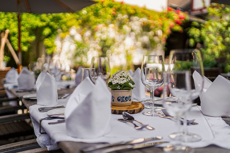 Mesa de jantar ao ar livre decorada com toalha branca, taças, talheres e flores, ideal para eventos ou celebrações ao ar livre.