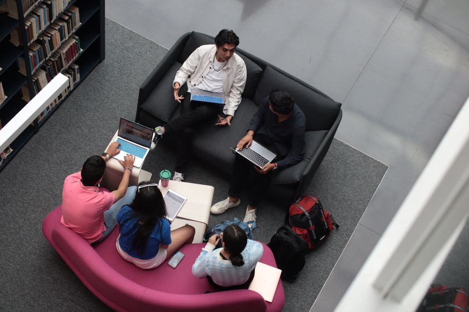 Grupo de estudantes ou profissionais discutindo em uma sala de estudos ou reunião, usando laptops e tablets.