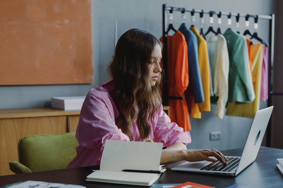 Mulher trabalhando em um laptop na rotina de gerenciamento de moda ou negócios de vestuário, com roupas coloridas ao fundo.