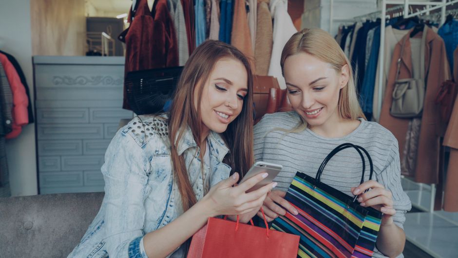 Duas jovens mulheres fazendo compras de roupas em uma loja, sorrindo e conversando enquanto olham o celular juntas.