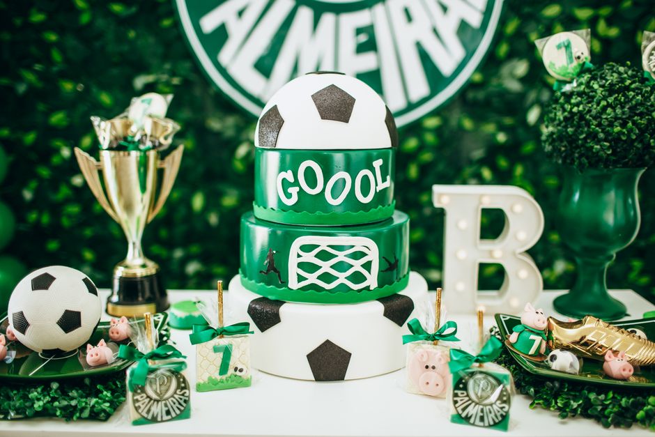 Mesa decorada com tema de futebol para festa infantil, incluindo bolo, troféu, enfeites e lembrancinhas em verde, branco e preto.