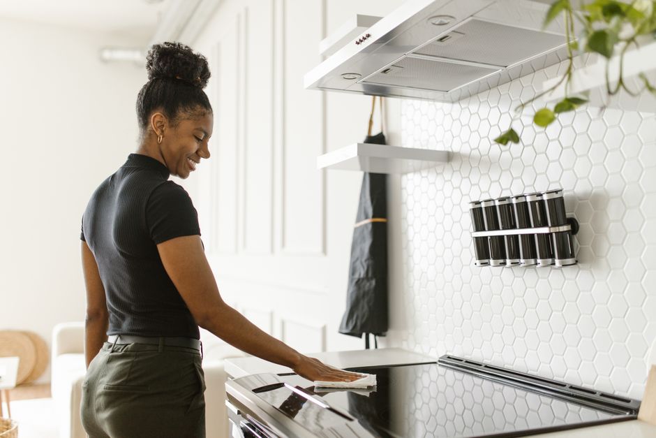 Mulher limpando a superfície de um fogão moderno em uma cozinha com decoração minimalista e paredes de azulejos hexagonais brancos.