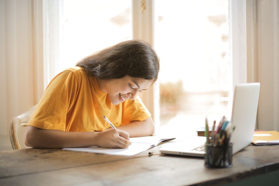 Mulher estudando e fazendo anotações em um ambiente de trabalho doméstico, com um laptop ao lado, simbolizando aprendizado ou pesquisa online.