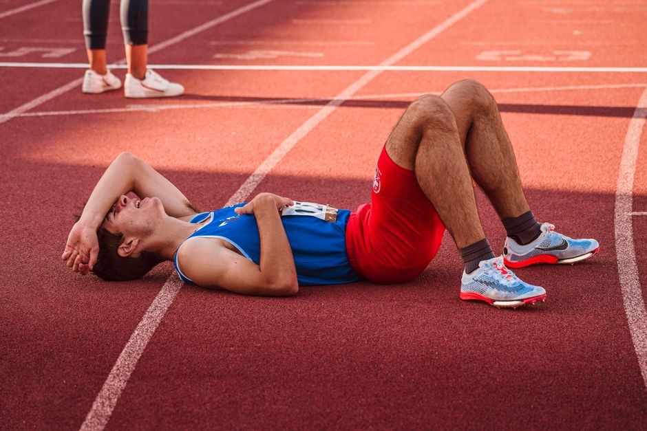 Atleta de corrida de velocidade deitado no chão de uma pista, aparentemente exausto após a prova, em um cenário esportivo ao ar livre.