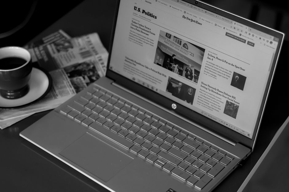 Imagem de um computador portátil exibindo uma página de notícias do The New York Times, acompanhado de uma xícara de café e jornais, sugerindo foco em notícias e leitura digital.