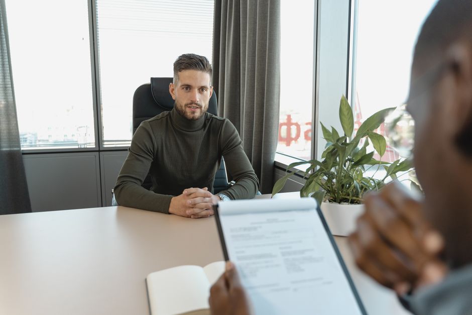 Homem realizando uma entrevista de emprego em ambiente de escritório com iluminação natural e plantas decorativas ao fundo.