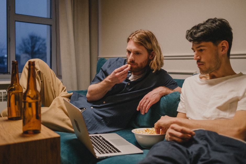 Dois jovens assistindo a um filme ou série em casa, relaxados no sofá com uma tigela de pipoca e uma laptop aberto, em um ambiente aconchegante na sala de estar.