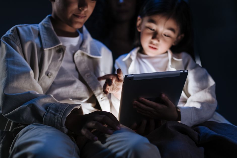 Crianças assistindo a conteúdo educativo em um tablet, em um ambiente escuro, evidenciando o uso de tecnologia na educação infantil.