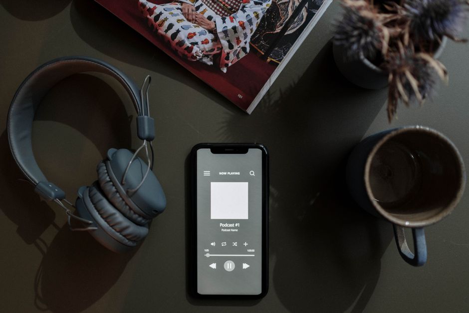 Imagem de uma mesa de trabalho com fone de ouvido, telefone com um podcast em reprodução, revista, roxinho com flores secas e duas canecas, direcionada a temas de consumo de conteúdo, rotina e produtividade.