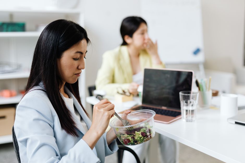 Duas mulheres em ambiente de trabalho, uma consumindo uma salada enquanto a outra aparenta estar pensativa ao lado de um laptop, em um escritório moderno.