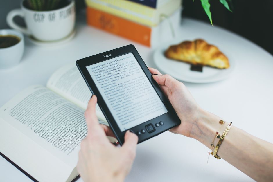 Pessoa lendo um livro digital em um ambiente de estudo ou trabalho, com café e croissants ao fundo, em uma mesa organizada.