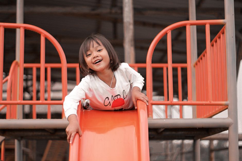 Menina sorridente brincando em um parque de diversões com escorregador laranja em um ambiente ao ar livre.