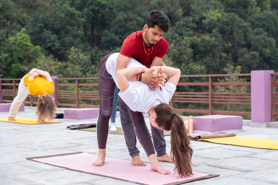Aula de yoga ao ar livre focada na prática de técnicas de relaxamento e alongamento, com instrutor auxiliando praticante em uma postura avançada.