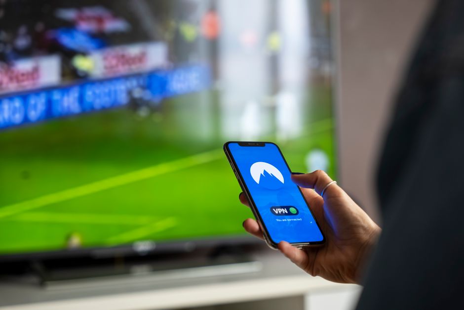Pessoa utilizando um smartphone com conexão VPN enquanto assiste a uma transmissão esportiva na TV, simbolizando conectividade e segurança digital.