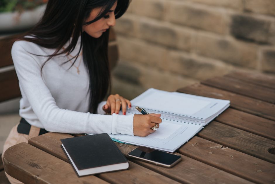 Mulher estudando ou planejando suas finanças pessoais ao ar livre, com materiais de estudo e tecnologia ao redor.