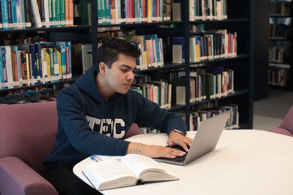 Jovem estudando ou realizando pesquisa em uma biblioteca, focado na tela do computador para um artigo de blog sobre pesquisa acadêmica ou estudo.
