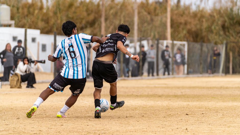 Dois jovens competindo em uma partida de futebol amador em um campo ao ar livre, com espectadores ao fundo.