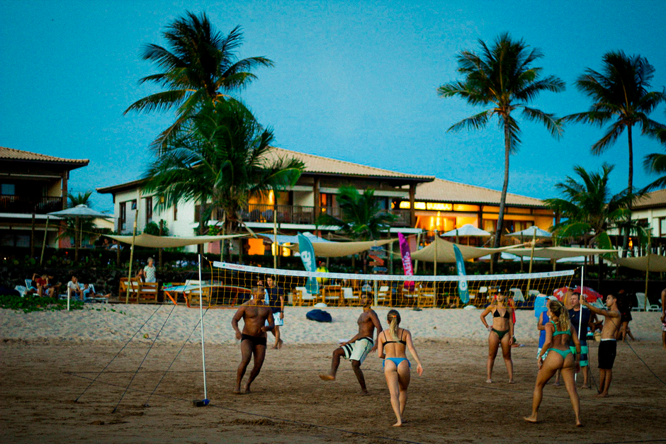 Imagem de uma partida de vôlei de praia ao pôr do sol, com pessoas jogando na areia e um resort ao fundo em clima de lazer e relaxamento.