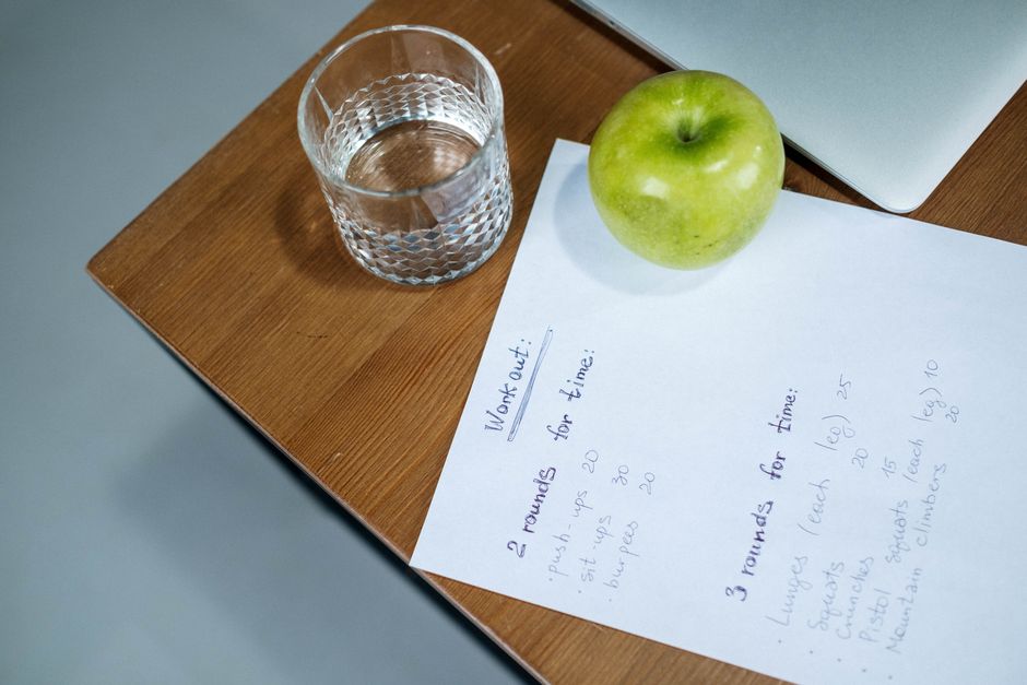 Imagem de uma mesa de trabalho com uma folha de anotações, uma maçã verde, um copo de água e um laptop, sugerindo foco em produtividade e organização pessoal.