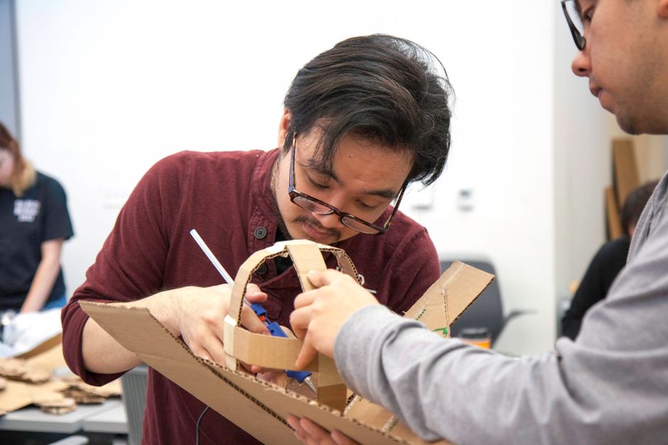 Homem ajustando um projeto artesanal de robô feito com materiais recicláveis, em ambiente de oficina ou sala de aula de tecnologia.