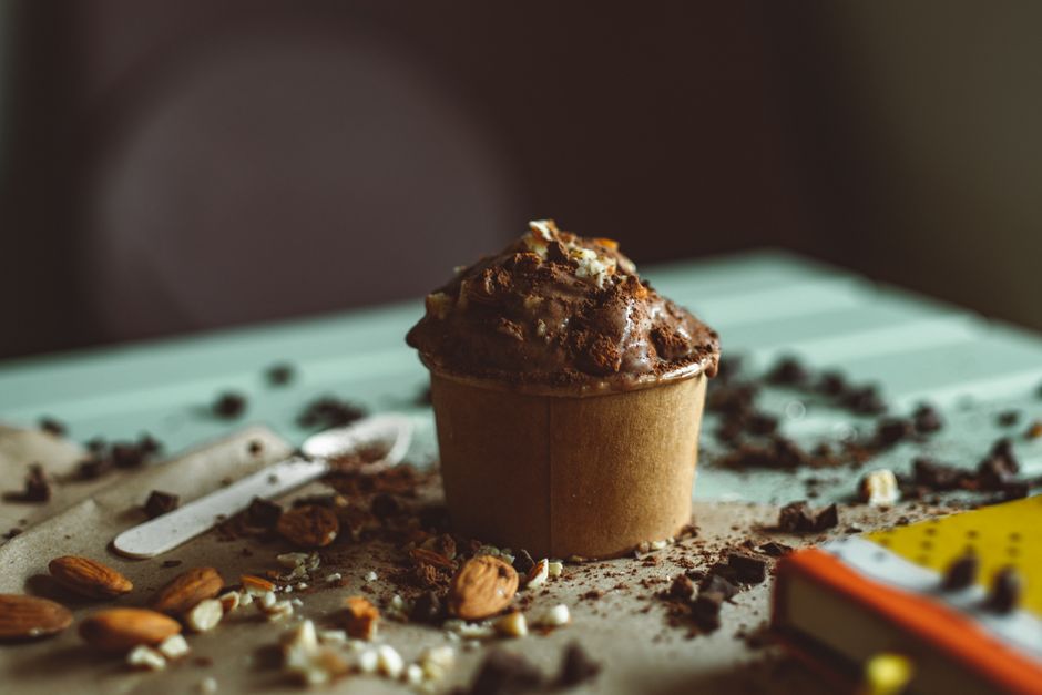 Imagem de um cupcake de chocolate sobre uma mesa decorada com pedaços de chocolate, castanhas e uma colher, criando uma atmosfera acolhedora e deliciosa.
