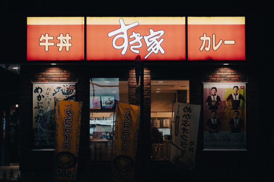 Imagem de fachada de restaurante japonês à noite, com sinalização iluminada e banners promocionais na entrada.