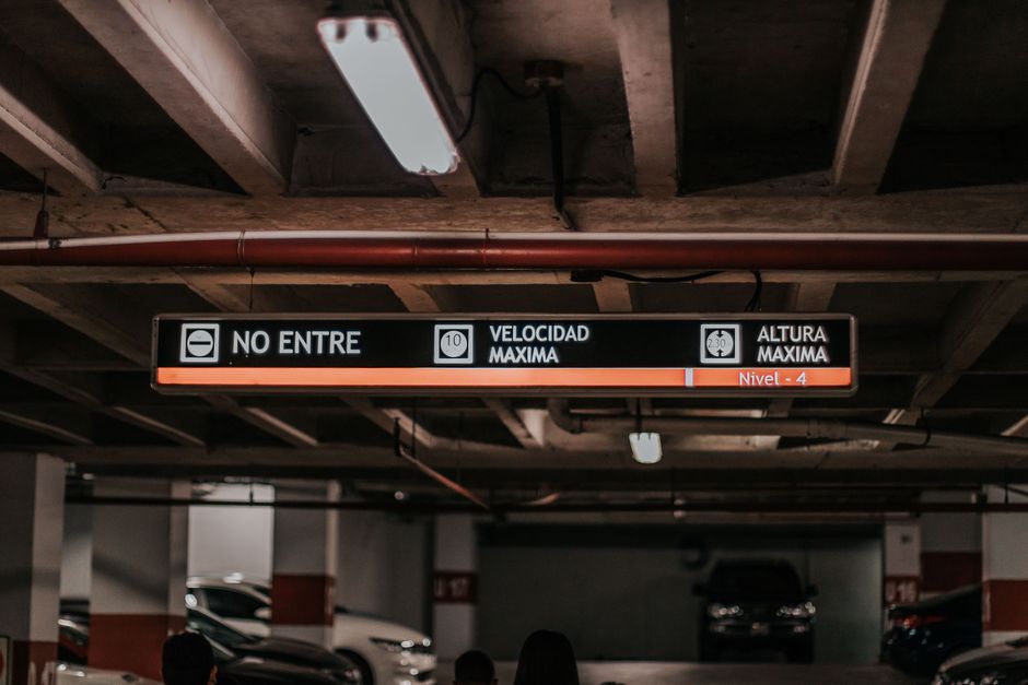 Placa de sinalização em estacionamento indicando limite de velocidade, altura máxima e nível de estacionamento, com iluminação de teto ao fundo.