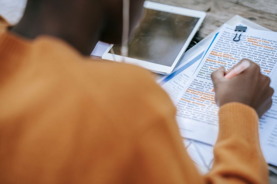 Pessoa estudando ou revisando documentos e anotações em um ambiente de trabalho ao ar livre, com tablet e materiais de leitura ao redor.