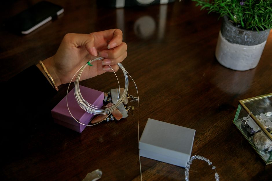 Pessoa segurando joias, possivelmente brincos, em uma mesa de madeira com vasos de plantas e caixas de acessórios ao redor, destacando o tema de acessórios e organização pessoal.
