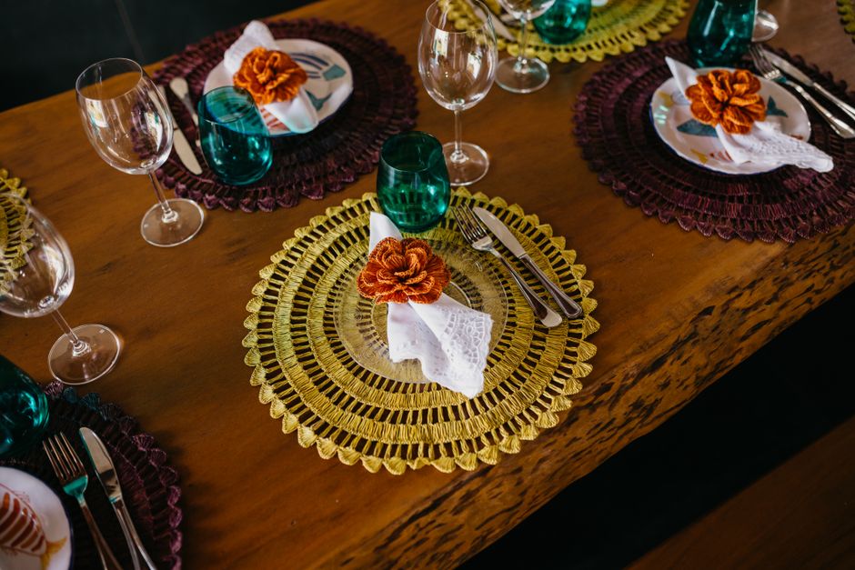Mesa de jantar decorada com toalhas de crochê coloridas e sousplats de palha, preparada para uma ocasião especial ou celebração em ambiente aconchegante.