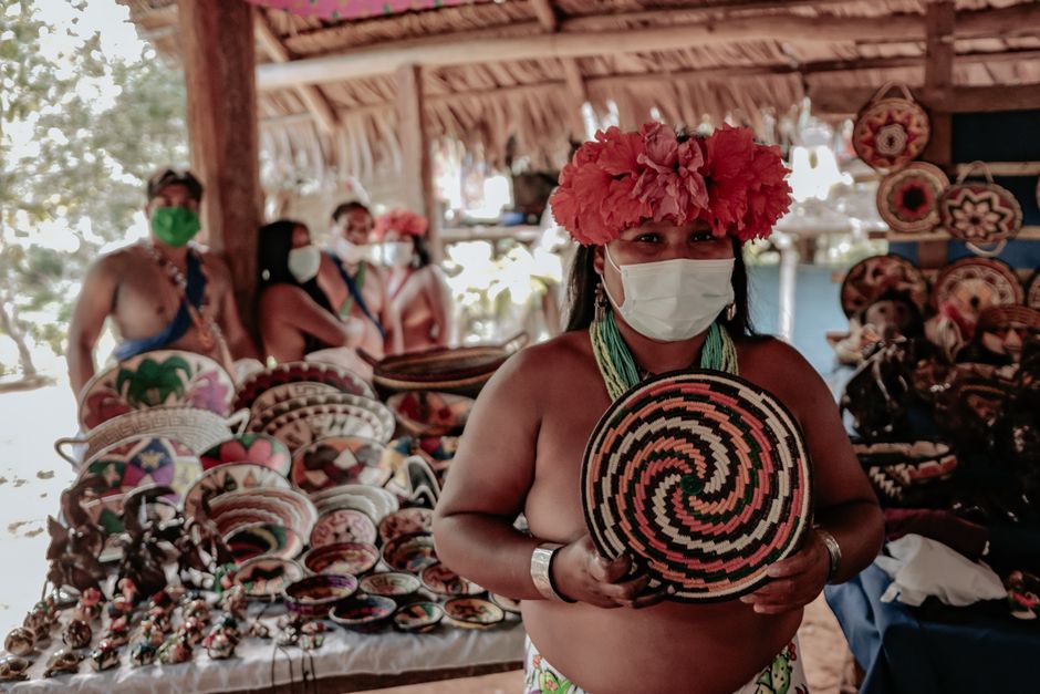 Mulher indígena em feira artesanal com acessórios tradicionais e produtos de artesanato, usando máscara facial para proteção, representando cultura e sustentabilidade.