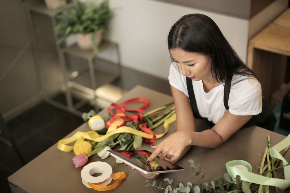 Mulher decoradora organizando flores e fitas coloridas para arranjo floral em ambiente interno aconchegante.