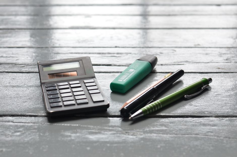 Imagem de uma atmosfera de estudo ou planejamento financeiro ao mostrar uma calculadora, corretor, e duas canetas sobre uma mesa de madeira escura.