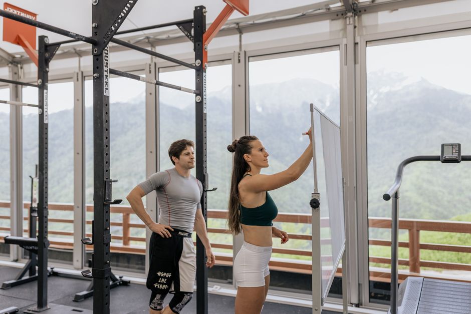 Mulher treinando musculação enquanto um instrutor observa ao fundo dentro de uma academia com vista montanhosa.