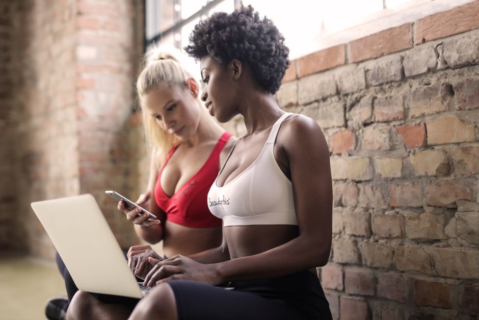 Duas mulheres em postura casual, uma usando top esportivo e a outra com blusa vermelha, interagindo com dispositivos móveis em um ambiente com parede de tijolos expostos, sugerindo um momento de socialização ou trabalho informal.