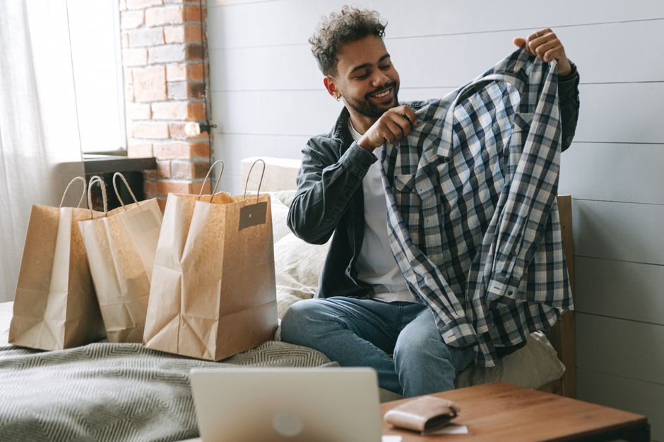 Homem feliz analisando roupas novas, com sacolas de compras ao lado, representando consumo e lifestyle urbano.