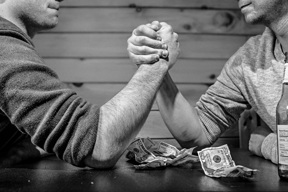 Duas pessoas envolvidas em um braço de ferro, simbolizando competitividade ou negociação, com dinheiro espalhado na mesa ao redor.