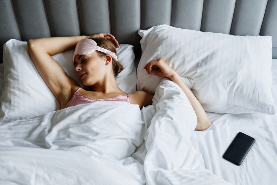 Mulher dormindo tranquilamente na cama, após um episódio de doença ou mal-estar, com uma máscara de dormir na cabeça e o celular ao lado.