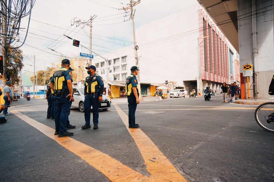 Imagem de policiais de trânsito coordenando o fluxo de veículos em uma esquina movimentada urbana, durante o dia.