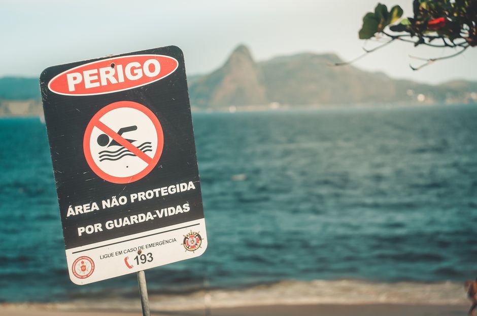 Placa de aviso de perigo em uma área aquática não protegida, alertando sobre riscos de segurança e a necessidade de cuidado ao nadar ou liberar vidas.