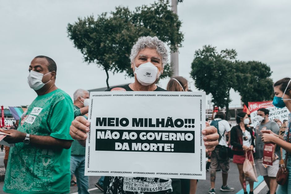 Mulher de cabelos grisalhos participando de protesto com cartaz contra a morte e o governo, em ambiente ao ar livre com várias pessoas ao fundo.