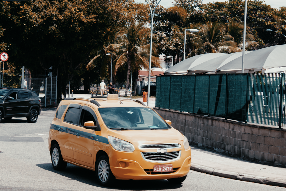Imagem de um carro de táxi amarelo estacionado em uma área urbana com árvores ao fundo, representando transporte público e mobilidade urbana.