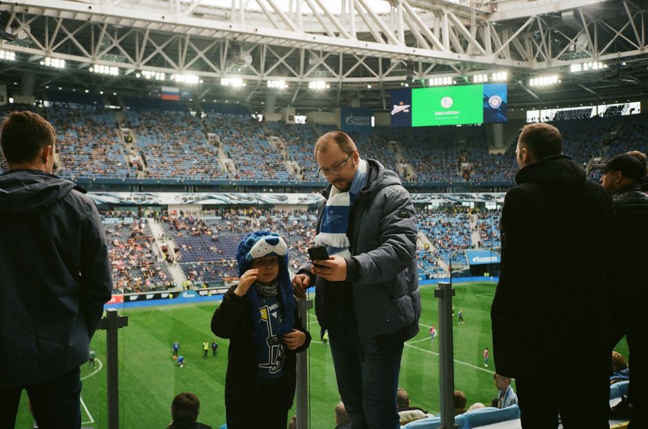 Família assistindo a uma partida de futebol em um estádio coberto, com o filho usando óculos de realidade virtual.
