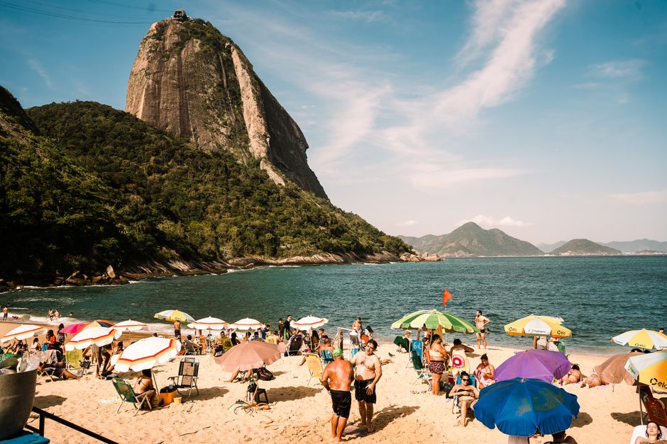 Praia com turistas sob guarda-sóis coloridos, com o famoso Monte do Pão de Açúcar ao fundo, em um dia ensolarado no Rio de Janeiro.