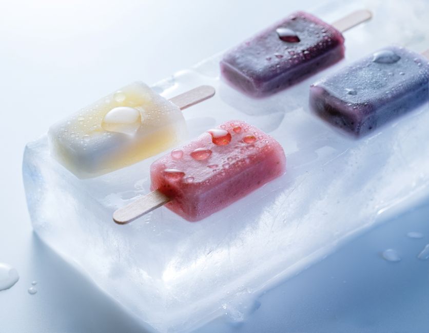 Três picolés coloridos em diferentes sabores repousando sobre um bloco de gelo, com gotas de água e sensação de frescor.