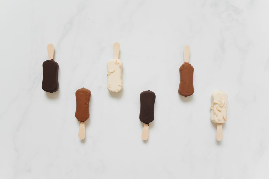 Imagem de cinco picolés de sabores diferentes dispostos sobre uma superfície de mármore branco, destacando variedade e cores variadas.