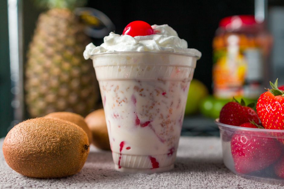 Foto de um copo de sobremesa com chantilly e cereja, com frutas variadas ao fundo, em um ambiente ao ar livre.