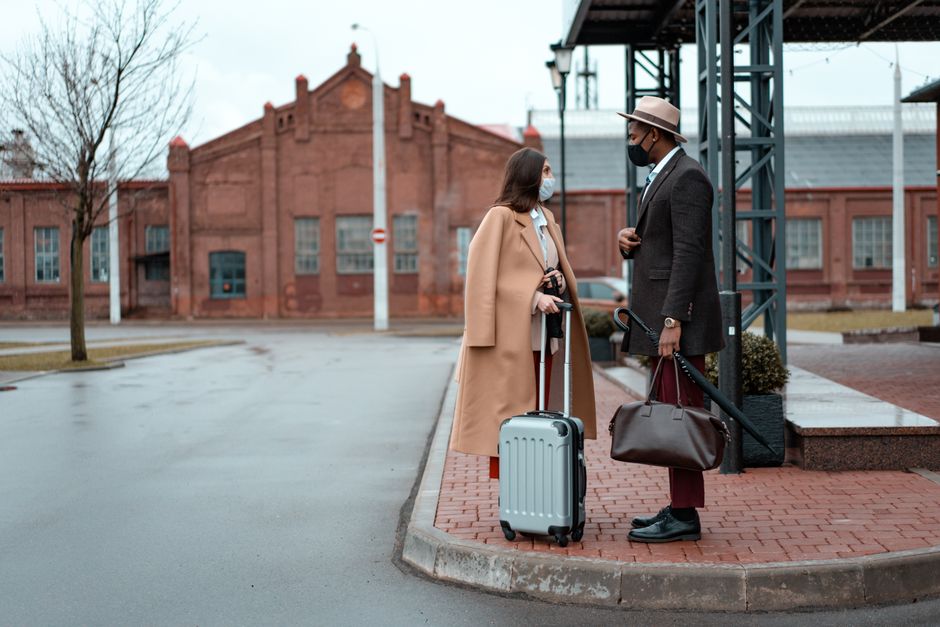Casal de viajantes conversando na área de embarque de uma estação, prontos para uma viagem de trem ou ônibus, com malas ao lado.