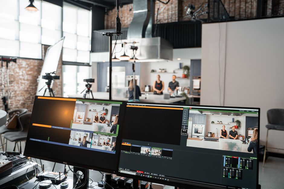 Ambiente de gravação de vídeo em estúdio com duas câmeras enfocando apresentadores na cozinha, com equipamentos de produção e editores ao fundo.