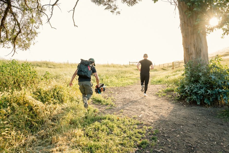 Duas pessoas caminhando por uma trilha ao ar livre durante o pôr do sol, com uma câmera profissional em mãos, em um ambiente natural e tranquilo.